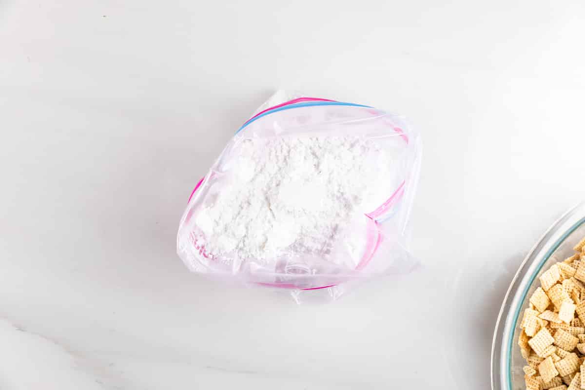 A resealable plastic bag filled with white powdered sugar rests on a marble surface. Part of a bowl with cereal pieces is visible on the right side of the image.