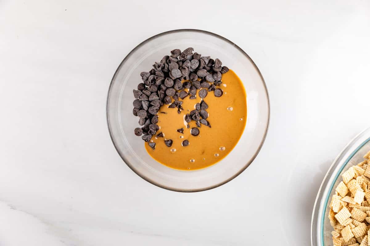 A glass bowl containing melted peanut butter and chocolate chips on a white surface. A portion of cereal is visible in the corner on a separate plate.