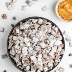 A bowl of puppy chow snack mix dusted with powdered sugar is on a white surface. Nearby are bowls of chocolate chips and peanut butter. A few chocolate chips and pieces of snack mix are scattered around. A striped napkin is partially visible.