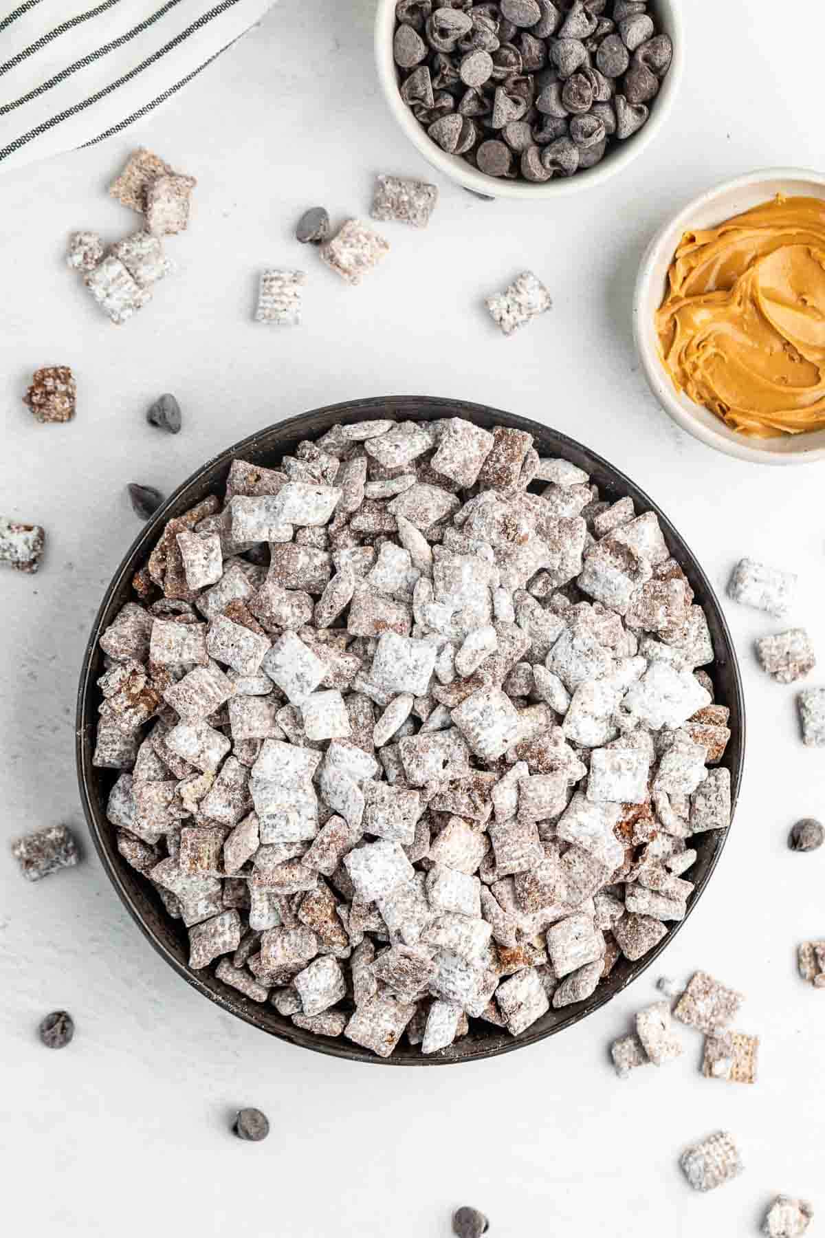 A bowl of puppy chow snack mix dusted with powdered sugar is on a white surface. Nearby are bowls of chocolate chips and peanut butter. A few chocolate chips and pieces of snack mix are scattered around. A striped napkin is partially visible.