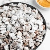 A bowl filled with Puppy Chow, a snack made from cereal coated in chocolate, peanut butter, and powdered sugar. Nearby are bowls containing peanut butter and chocolate chips. The surface is white, contrasting with the snacks texture.