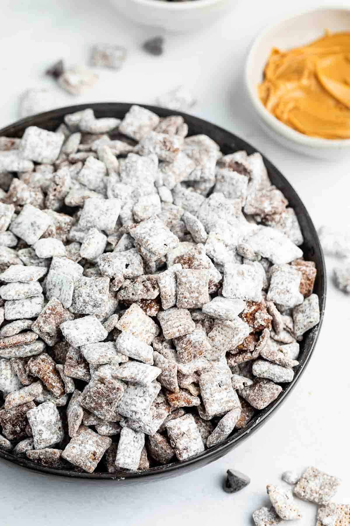 A bowl filled with Puppy Chow, a snack made from cereal coated in chocolate, peanut butter, and powdered sugar. Nearby are bowls containing peanut butter and chocolate chips. The surface is white, contrasting with the snacks texture.
