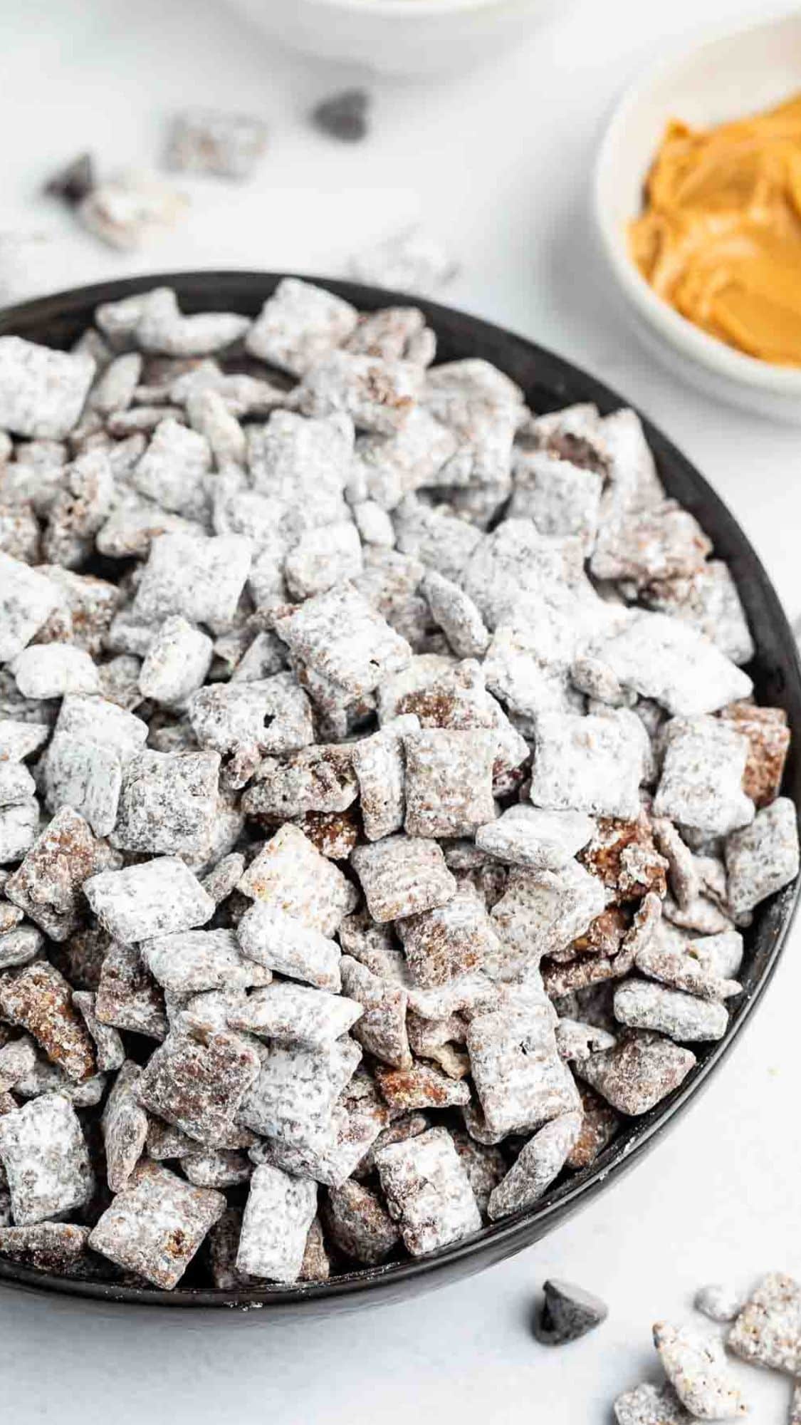 A black bowl filled with powdered sugar-coated snack pieces, likely a mix of cereal and chocolate, sits on a white surface. Nearby are small bowls with peanut butter and chocolate chips, suggesting ingredients for the snack.