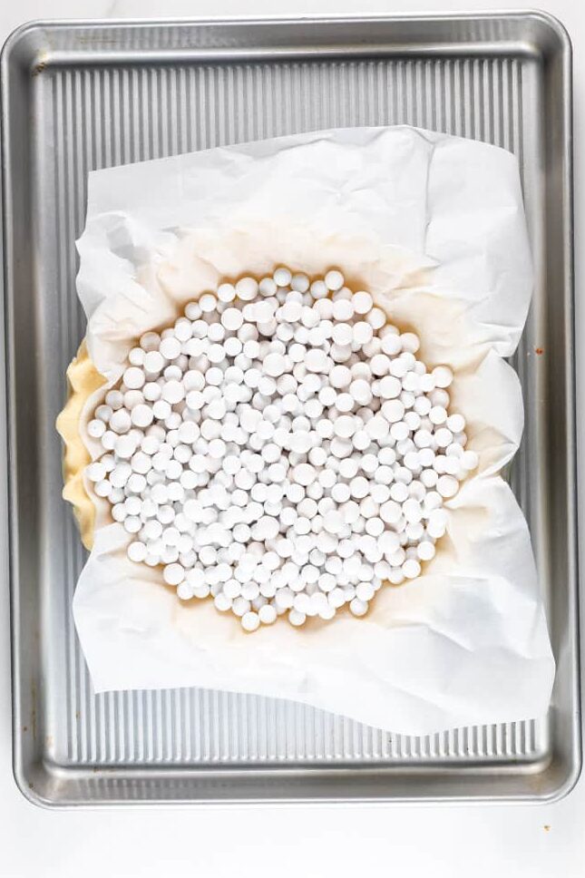 A pie crust is lined with parchment paper and filled with white baking weights, placed on a silver baking sheet.