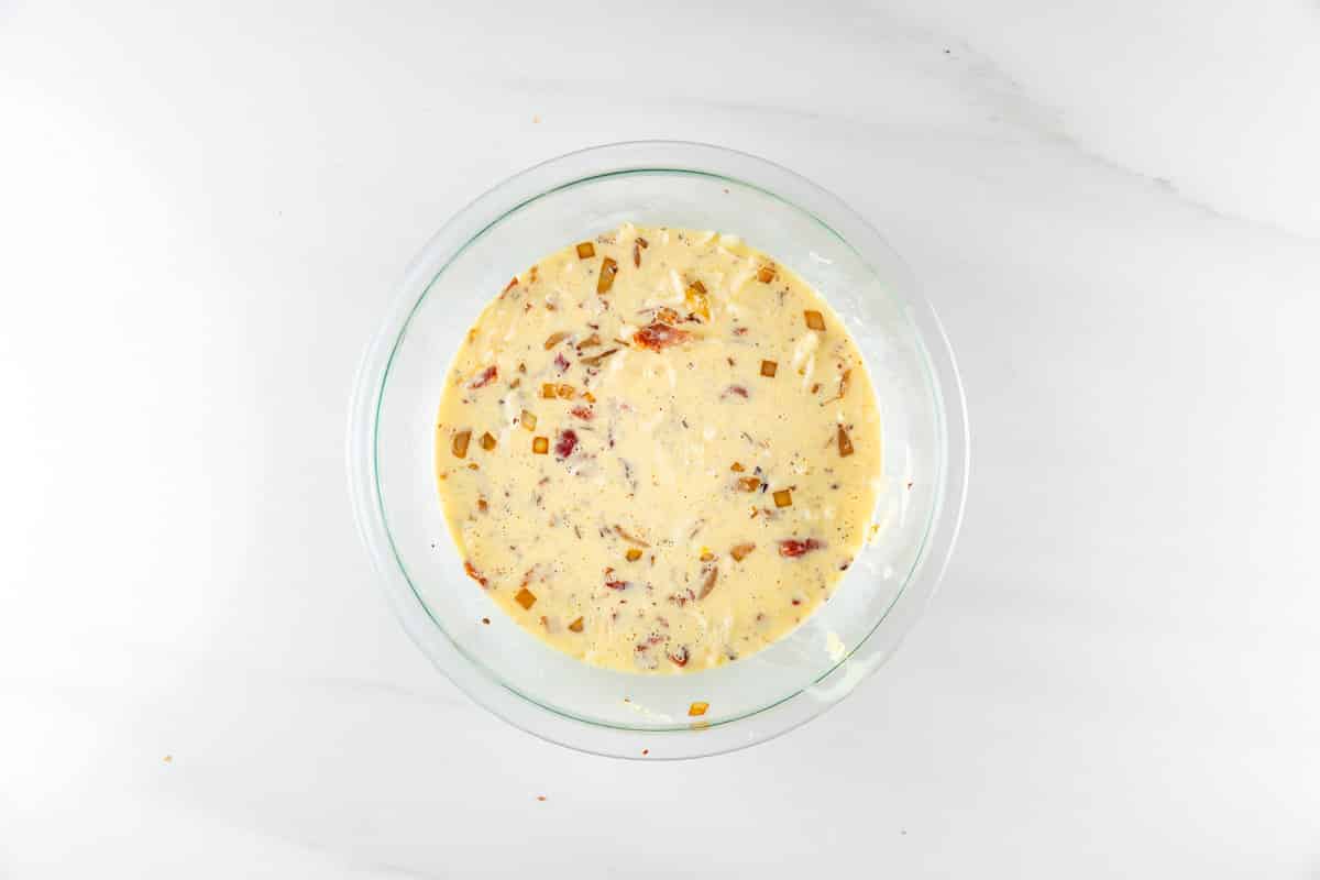 A glass bowl containing a creamy mixture with chopped pieces of dried fruit on a white marble surface.