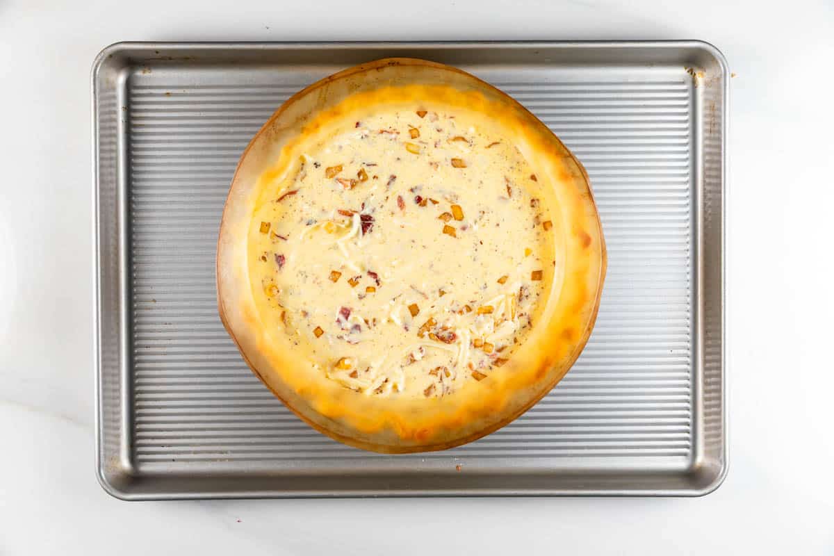 A round pumpkin filled with a creamy mixture sits in the center of a silver baking pan. The pumpkin has a toasted, golden-brown edge and contains a filling with visible chunks of onion and herbs.