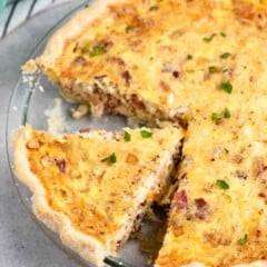 A freshly baked quiche with a slice cut out, revealing a flaky crust and a creamy filling with bits of bacon and herbs. The dish is garnished with chopped parsley. The quiche is placed on a gray countertop beside a striped cloth and eggshells.