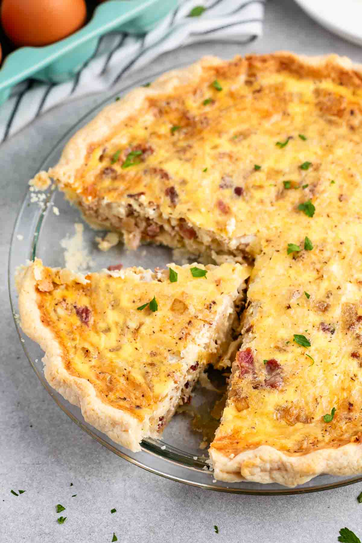 A freshly baked quiche with a slice cut out, revealing a flaky crust and a creamy filling with bits of bacon and herbs. The dish is garnished with chopped parsley. The quiche is placed on a gray countertop beside a striped cloth and eggshells.