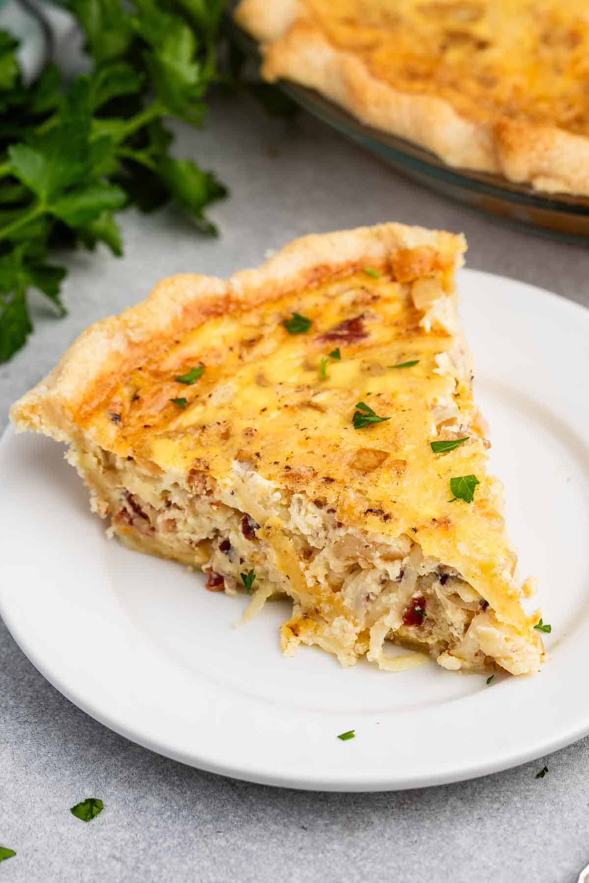 A slice of quiche on a white plate. The quiche has a golden crust and is filled with a creamy mixture containing small bits of bacon and herbs. Fresh parsley is scattered on top and around the plate.