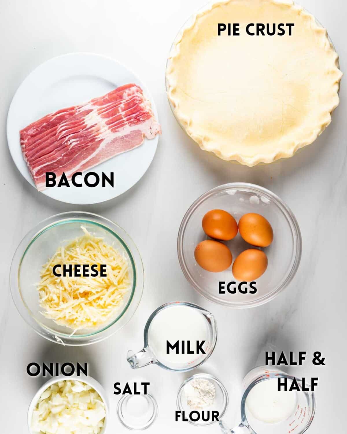 Ingredients for a quiche are arranged on a white surface. Items include a pie crust, bacon, shredded cheese, whole eggs, chopped onion, salt, flour, milk, and half & half. Each is labeled with black text.