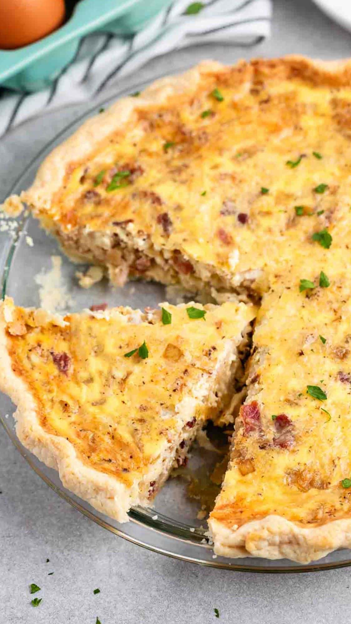 A sliced quiche in a glass pie dish, with a golden crust and savory filling containing visible pieces of bacon and herbs. One slice is slightly pulled out. Its placed on a gray surface with a striped cloth and eggs in the background.