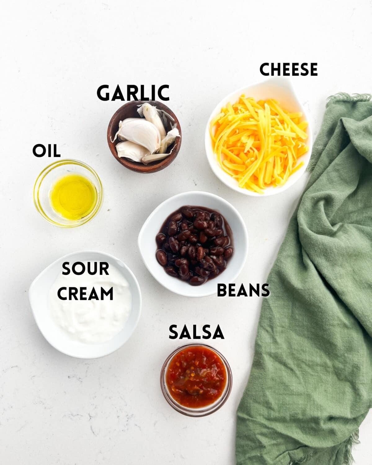 Ingredients arranged on a white surface: garlic cloves, shredded cheese, a green cloth, olive oil in a small dish, black beans in a bowl, sour cream, and salsa in separate bowls. Labels identify each ingredient.