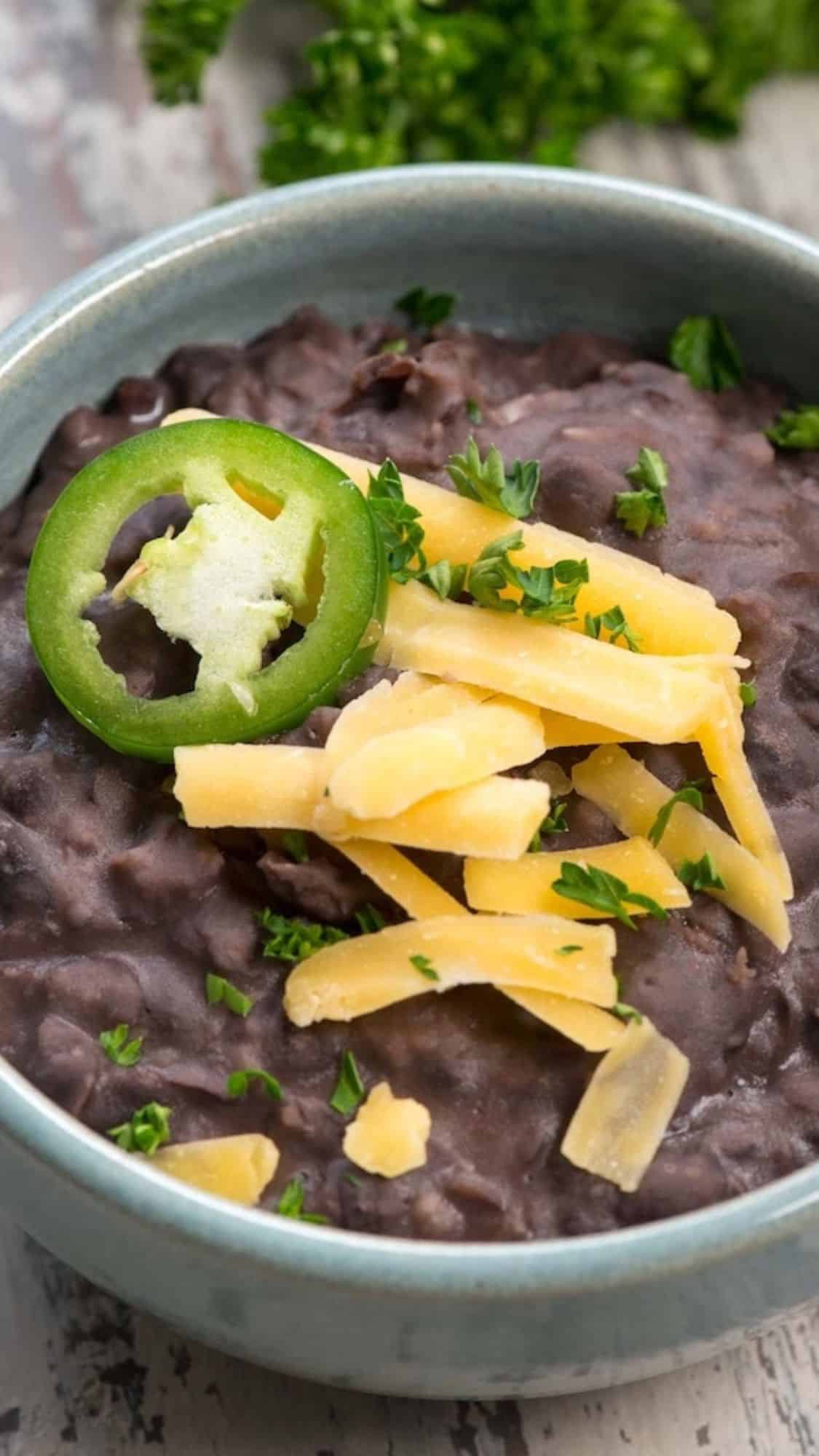 A bowl of black bean soup topped with shredded cheese and a slice of jalapeño. Fresh parsley is sprinkled on top. The bowl is placed on a rustic wooden surface with some greenery in the background.