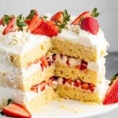 A sliced three-layer vanilla cake is topped with fresh strawberries and white frosting. Each layer contains cream and strawberry pieces. The cake is displayed on a white cake stand against a light gray background.