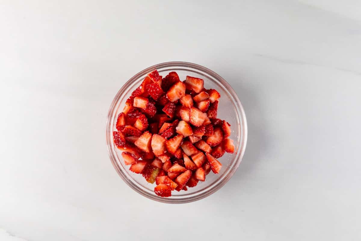 A glass bowl filled with small, evenly diced strawberries sits on a light gray surface. The vibrant red fruit contrasts against the neutral background, highlighting the strawberries freshness and texture.