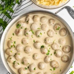 A pan filled with meatballs in a creamy white sauce, garnished with chopped parsley. Next to the pan is a bowl of uncooked egg noodles. A striped cloth and sprigs of fresh parsley are placed beside them.