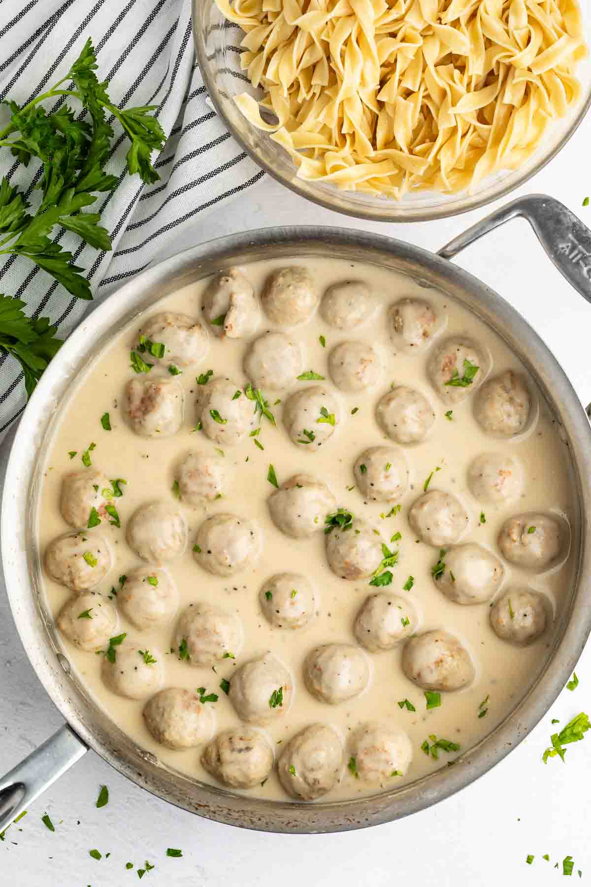 A pan filled with meatballs in a creamy white sauce, garnished with chopped parsley. Next to the pan is a bowl of uncooked egg noodles. A striped cloth and sprigs of fresh parsley are placed beside them.