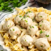 A plate of creamy pasta topped with meatballs in a white sauce, garnished with chopped herbs. A glass of red wine and fresh parsley are in the background.