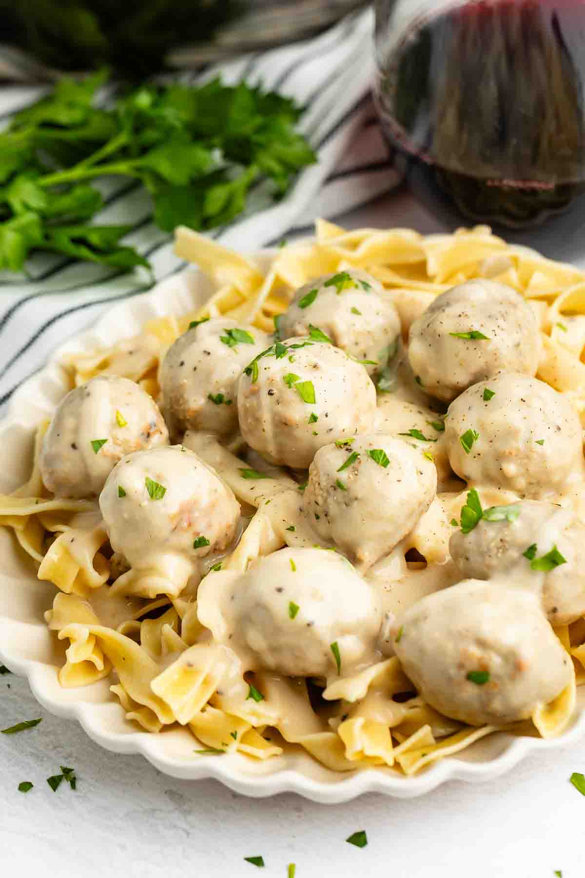 A plate of creamy pasta topped with meatballs in a white sauce, garnished with chopped herbs. A glass of red wine and fresh parsley are in the background.