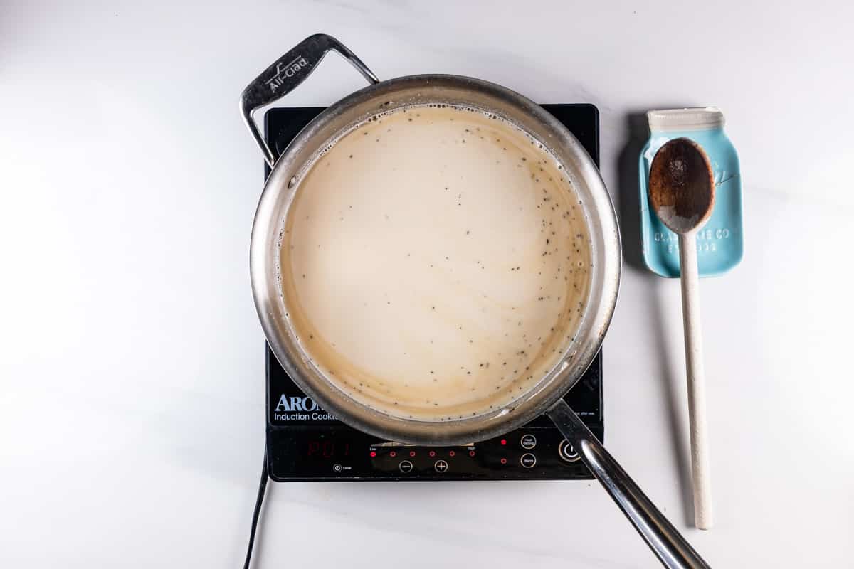 A pot of creamy soup with herbs on an induction cooktop. Next to it is a blue container labeled FARM FRESH MILK with a wooden spoon resting on top, all on a white marble countertop.