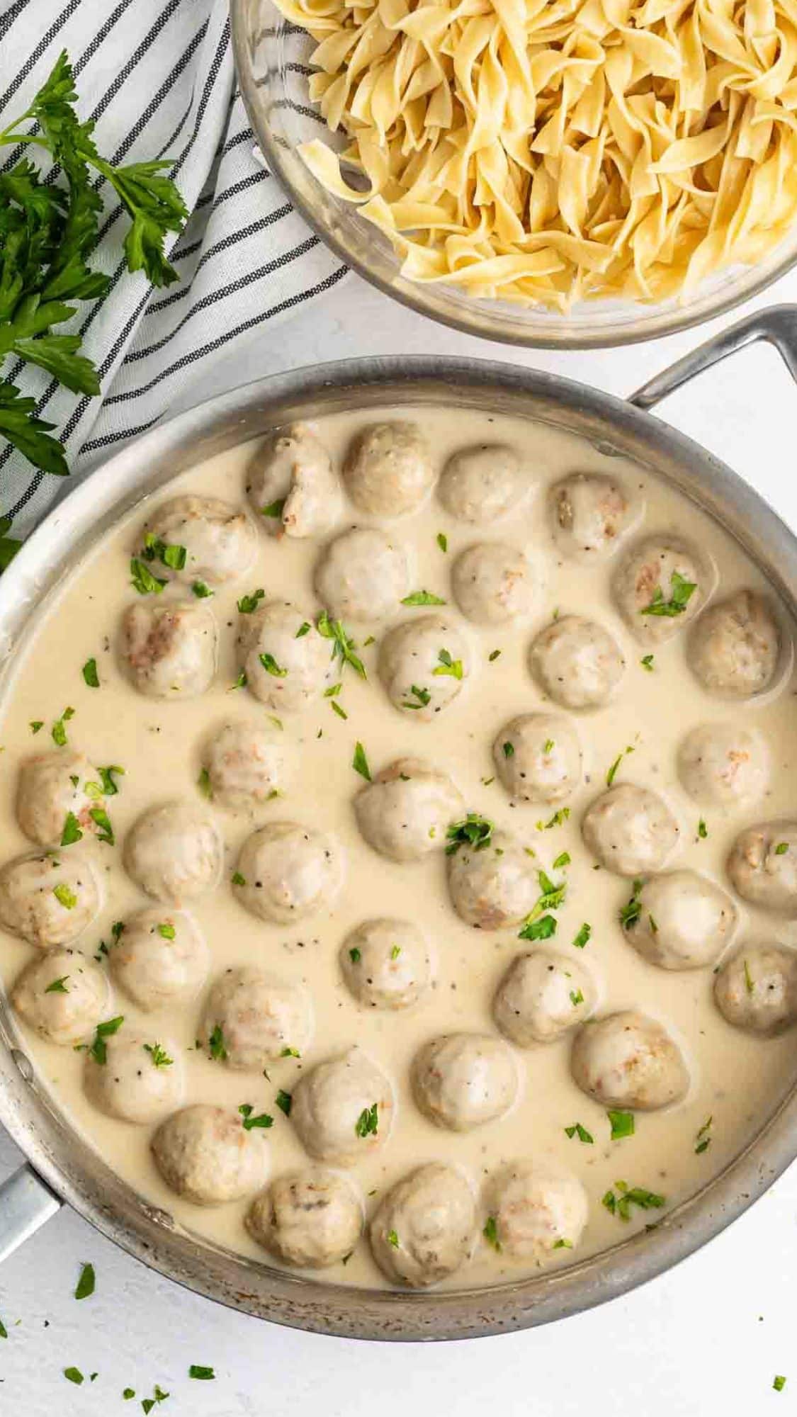 A skillet filled with meatballs in a creamy white sauce, garnished with chopped parsley. Next to it, a bowl of uncooked egg noodles. A striped cloth and some fresh parsley are visible in the background.