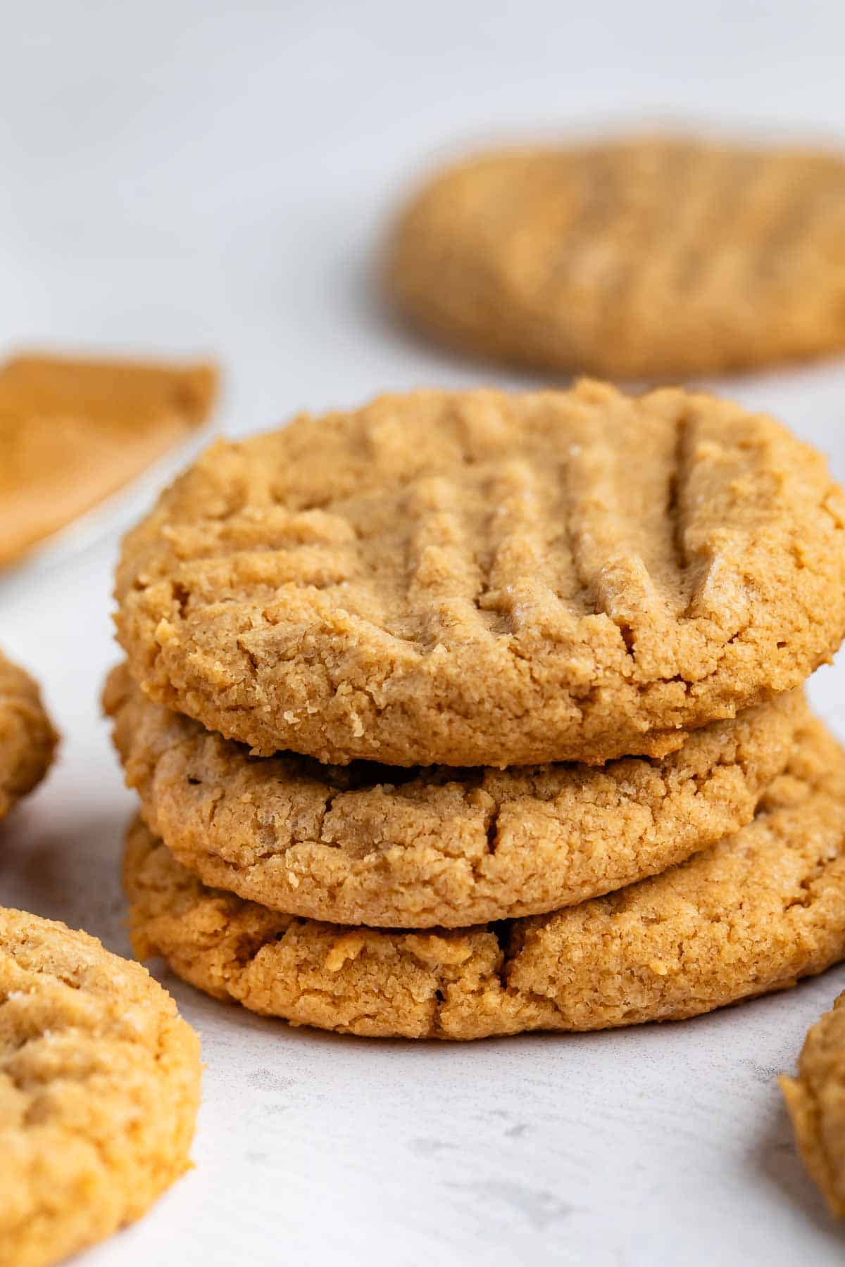 A stack of three peanut butter cookies with a crisscross fork pattern on top, surrounded by more cookies on a light surface.