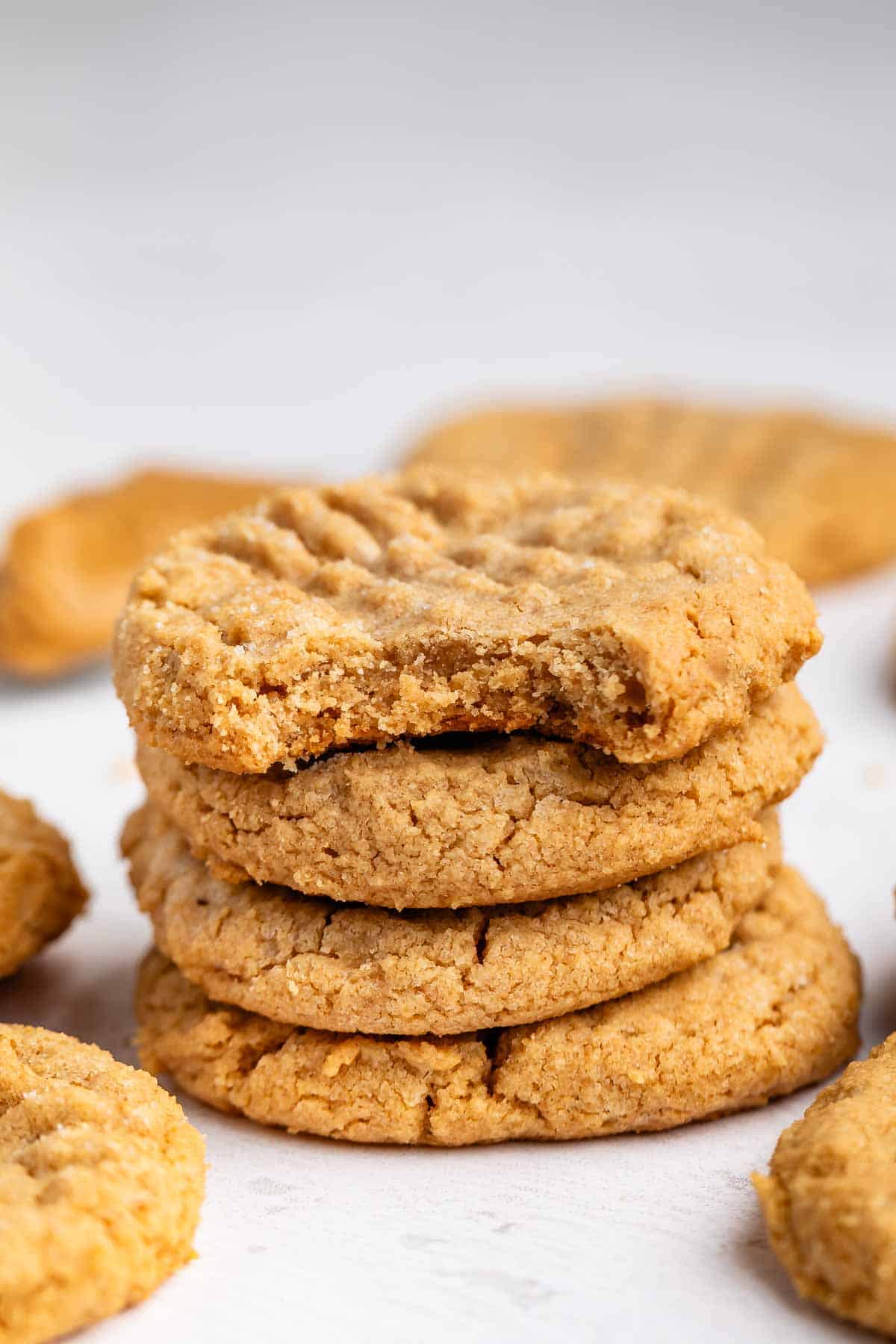 A stack of four peanut butter cookies sits on a white surface. The top cookie has a bite taken out of it, and more cookies are scattered in the background.