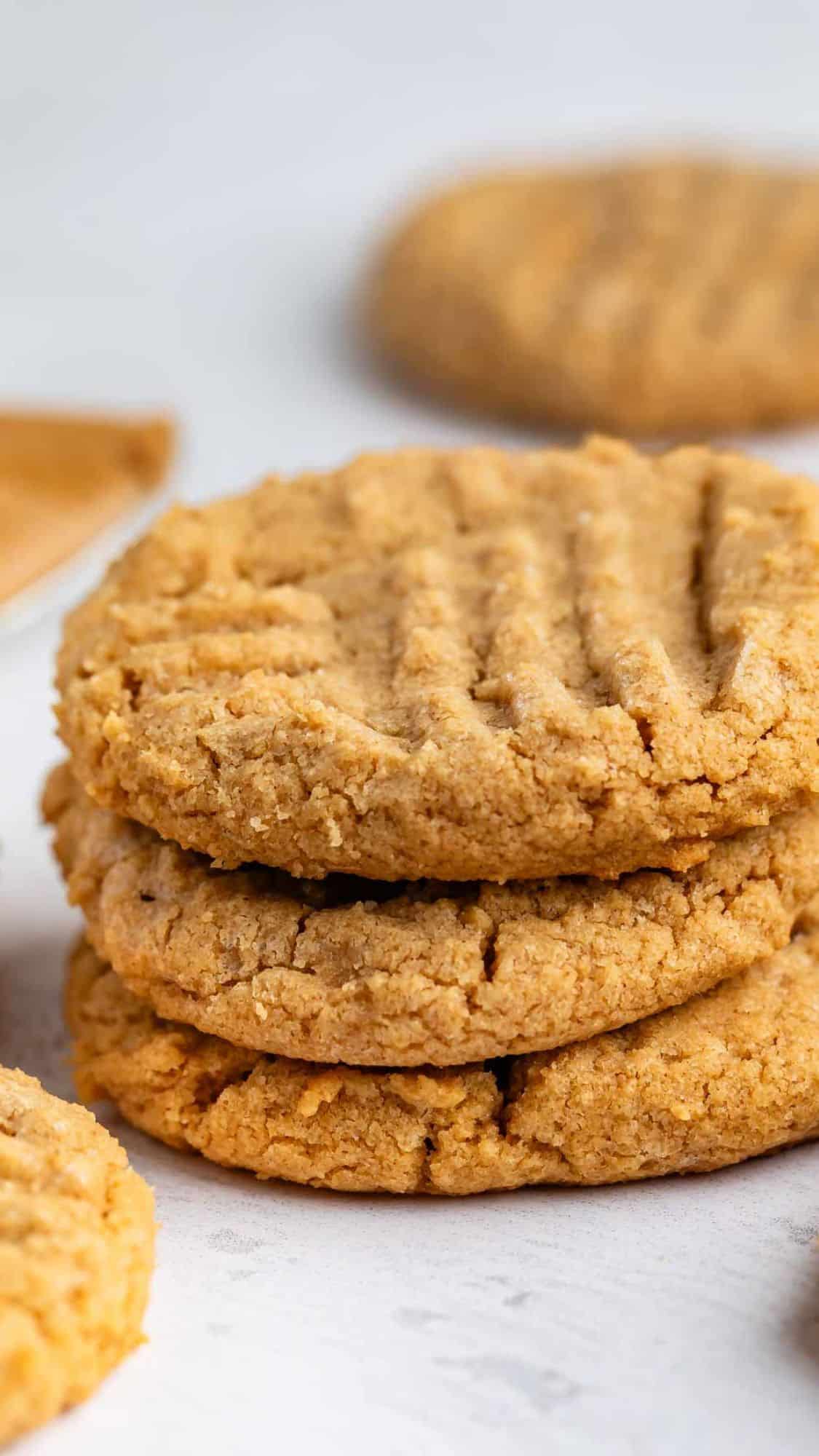 A stack of three golden brown peanut butter cookies with a crisscross fork pattern on top, sitting on a light surface with more cookies in the background.