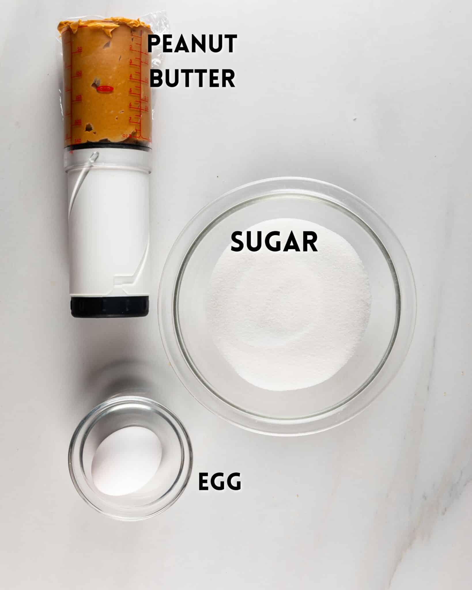 A measuring cup of peanut butter, a glass bowl of sugar, and a small glass bowl with one egg are arranged on a white surface. Each ingredient is labeled in bold black text.