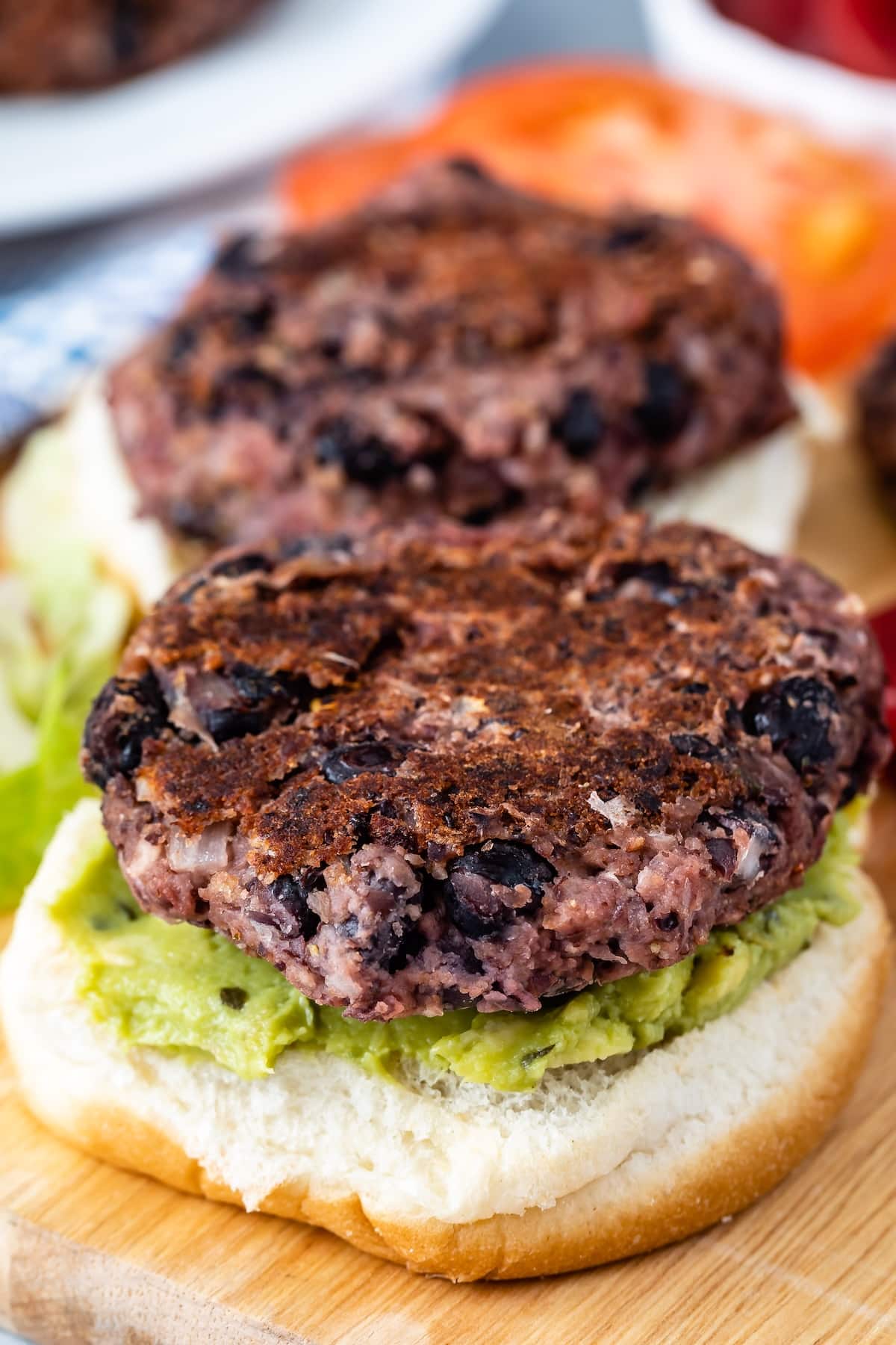 Close-up of a black bean burger on a wooden board. The burger patty is placed on a bun spread with guacamole, with visible lettuce underneath. The patty appears well-cooked and textured. In the background, there are blurred slices of tomato.