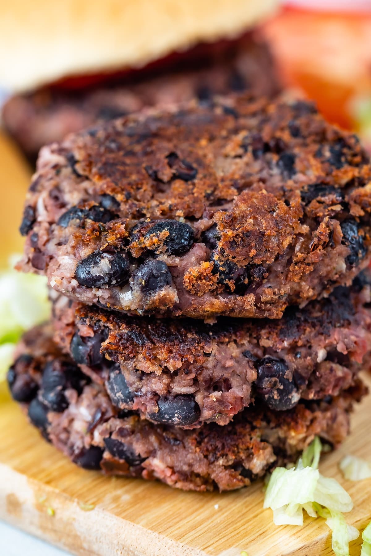 Close-up of three stacked black bean burgers with a crispy texture on a wooden surface. Lettuce is visible around the burgers, and a blurred bun with tomato slices can be seen in the background.