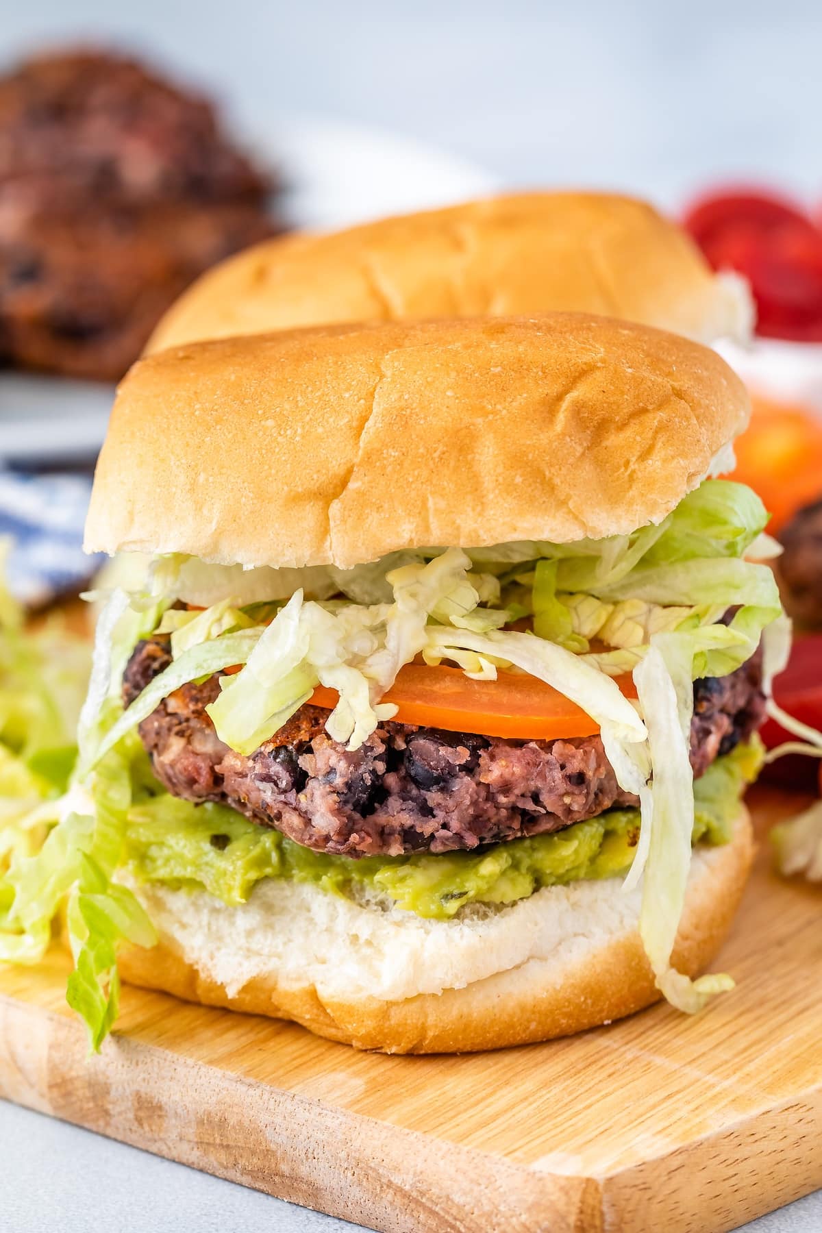 Veggie burger with a black bean patty, layered with fresh tomato slices, shredded lettuce, and guacamole, served in a soft bun on a wooden board. Blurred background shows additional ingredients and another burger.