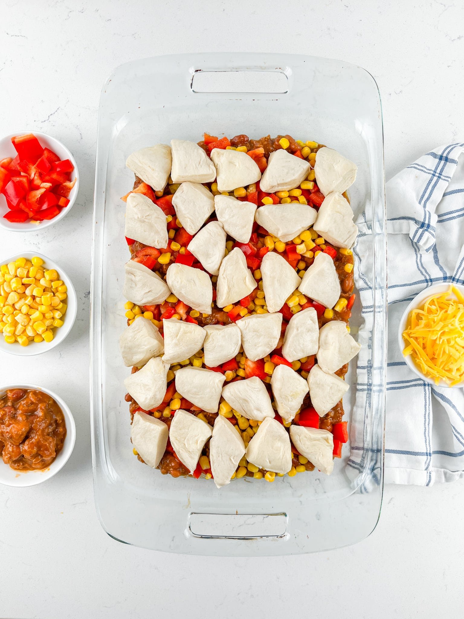 A glass baking dish filled with layered ingredients, topped with biscuit pieces, red bell peppers, corn, and ground meat. Surrounding the dish are small bowls of corn, diced red peppers, salsa, and shredded cheese on a white surface.