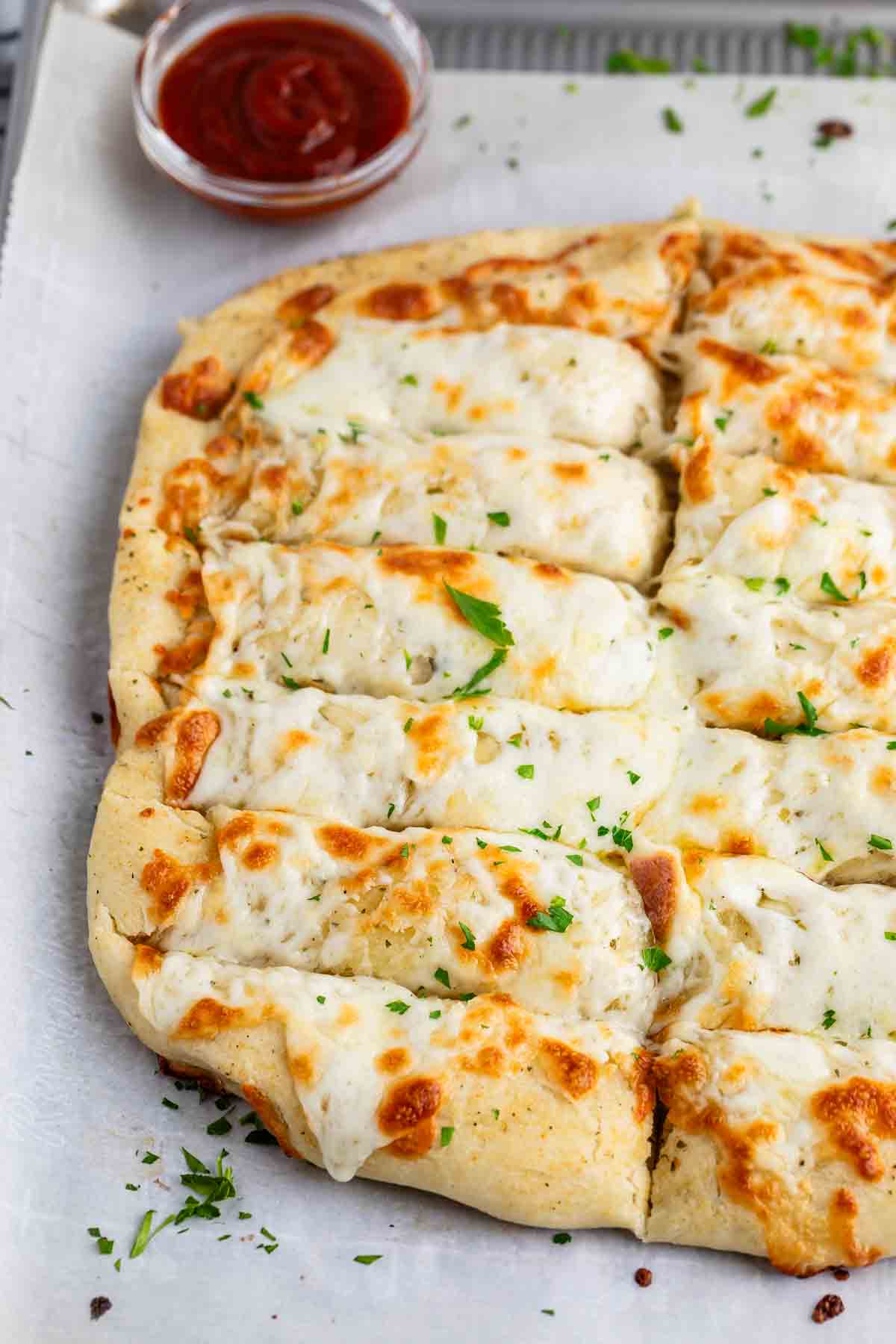 A rectangular cheesy bread topped with melted cheese and sprinkled with parsley is sliced into pieces on a baking sheet. A small bowl of marinara sauce is placed in the background.