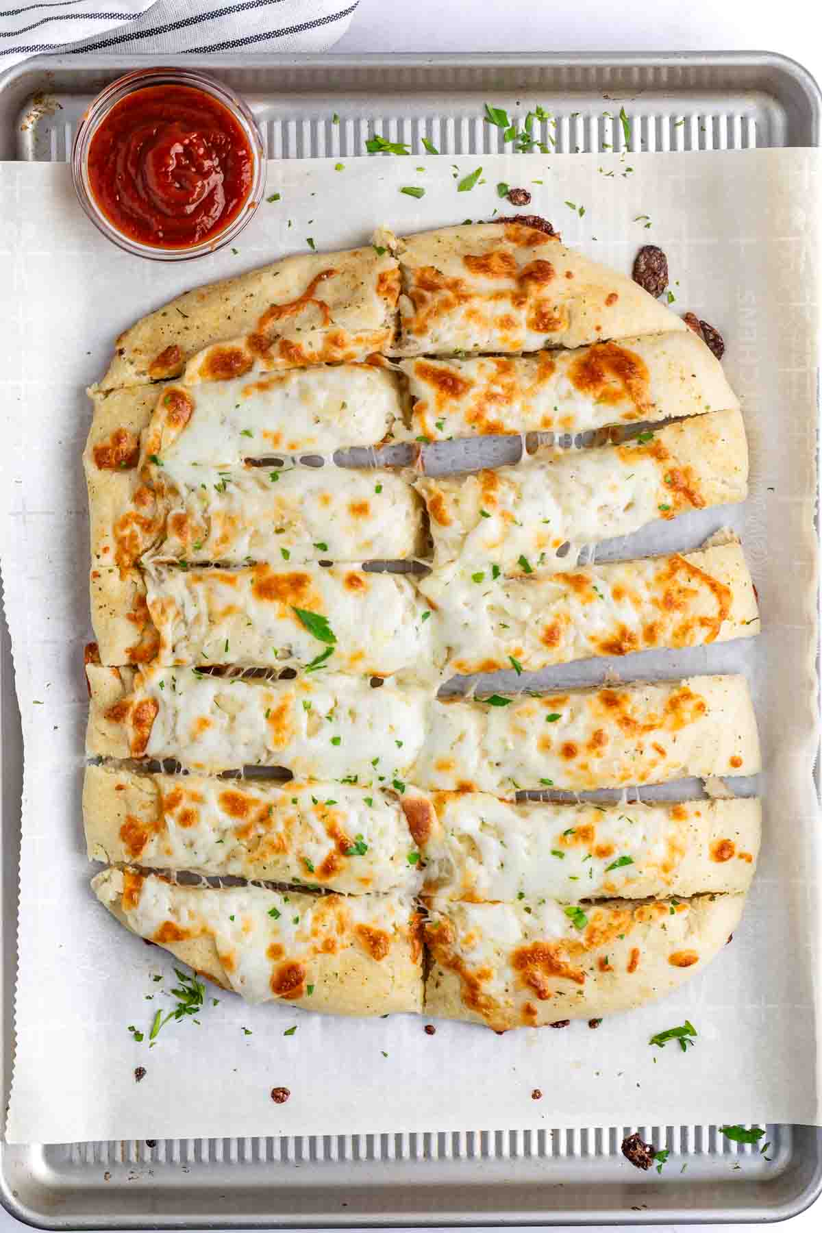 A rectangular cheese bread on a baking sheet lined with parchment paper, cut into strips. The bread is topped with melted cheese and garnished with parsley. A small cup of marinara sauce is on the side.