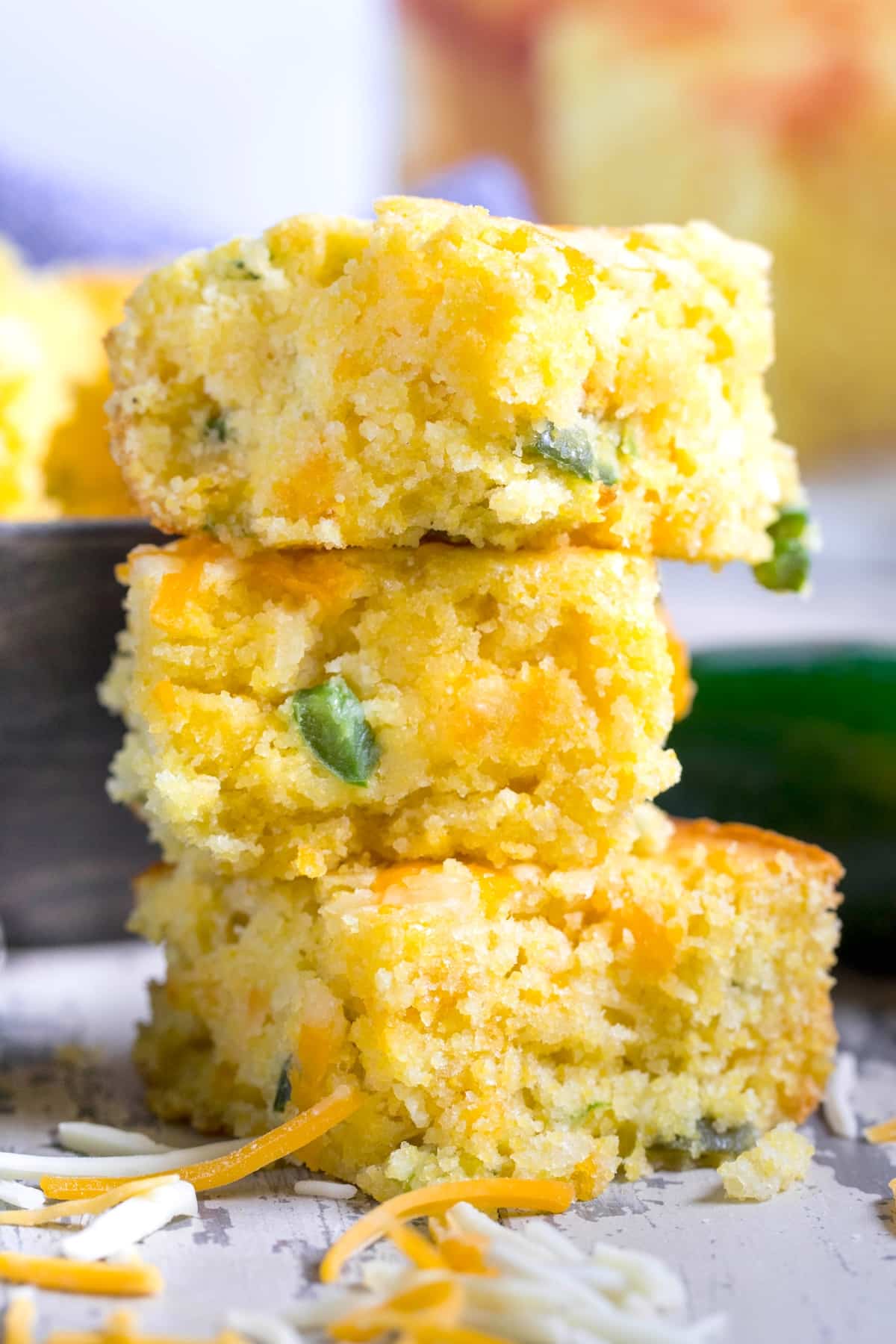 Three pieces of jalapeño cheddar cornbread stacked vertically. The bread appears moist with visible chunks of cheddar and bits of jalapeño, set on a rustic wooden surface with scattered shredded cheese and a blurred background.