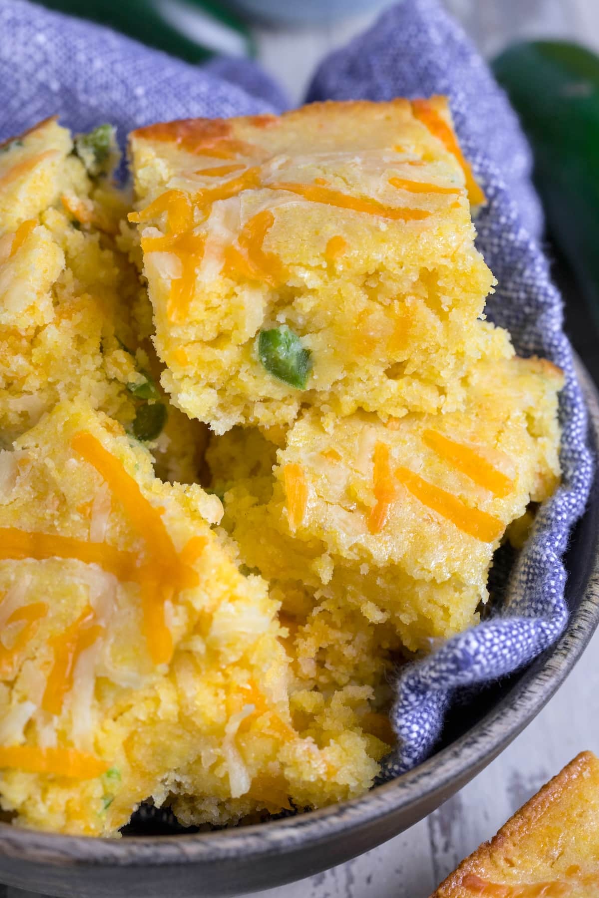 Close-up of a bowl containing pieces of cornbread topped with melted cheese. The cornbread is golden with visible green chili pieces, placed on a blue cloth napkin. The background has a rustic, wooden texture.