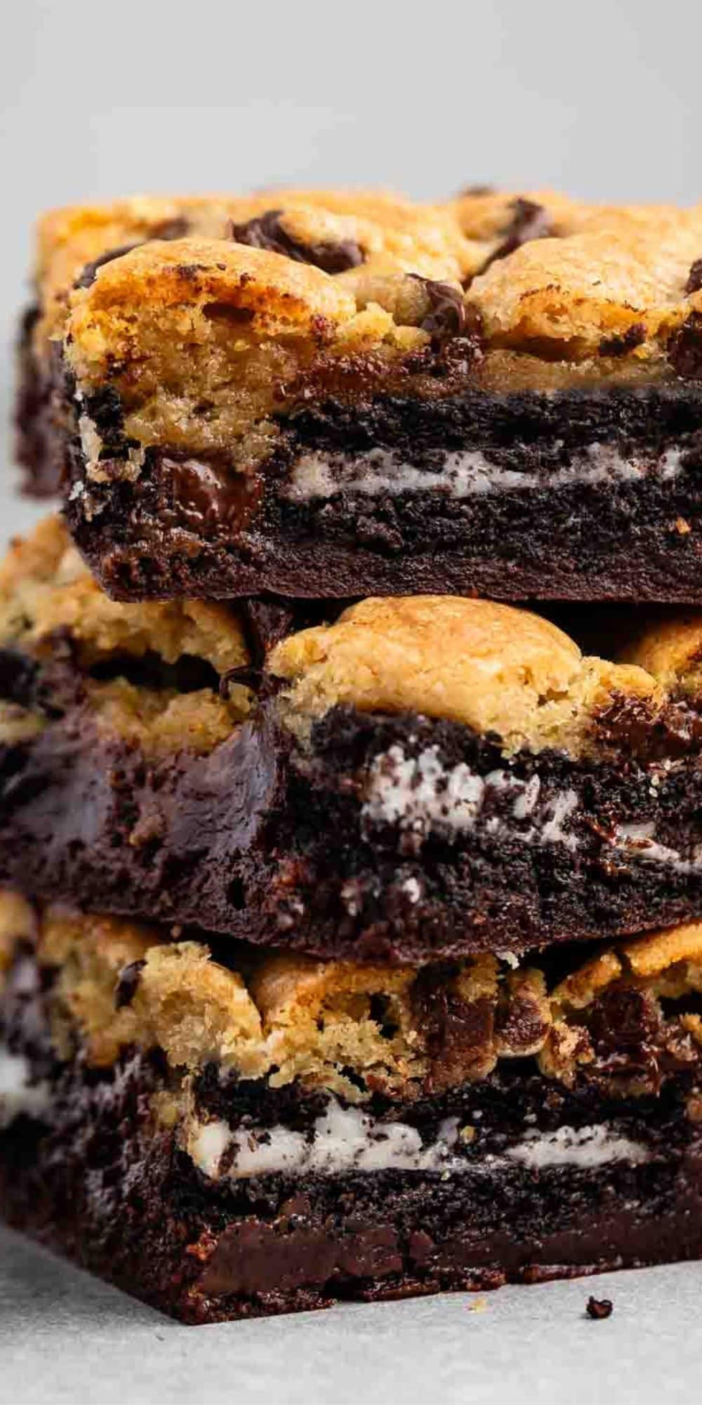 A close-up of a stack of dessert bars with layers of chocolate chip cookie, chocolate sandwich cookies, and brownie. The bars have a gooey texture with visible chocolate chunks and cookie pieces.