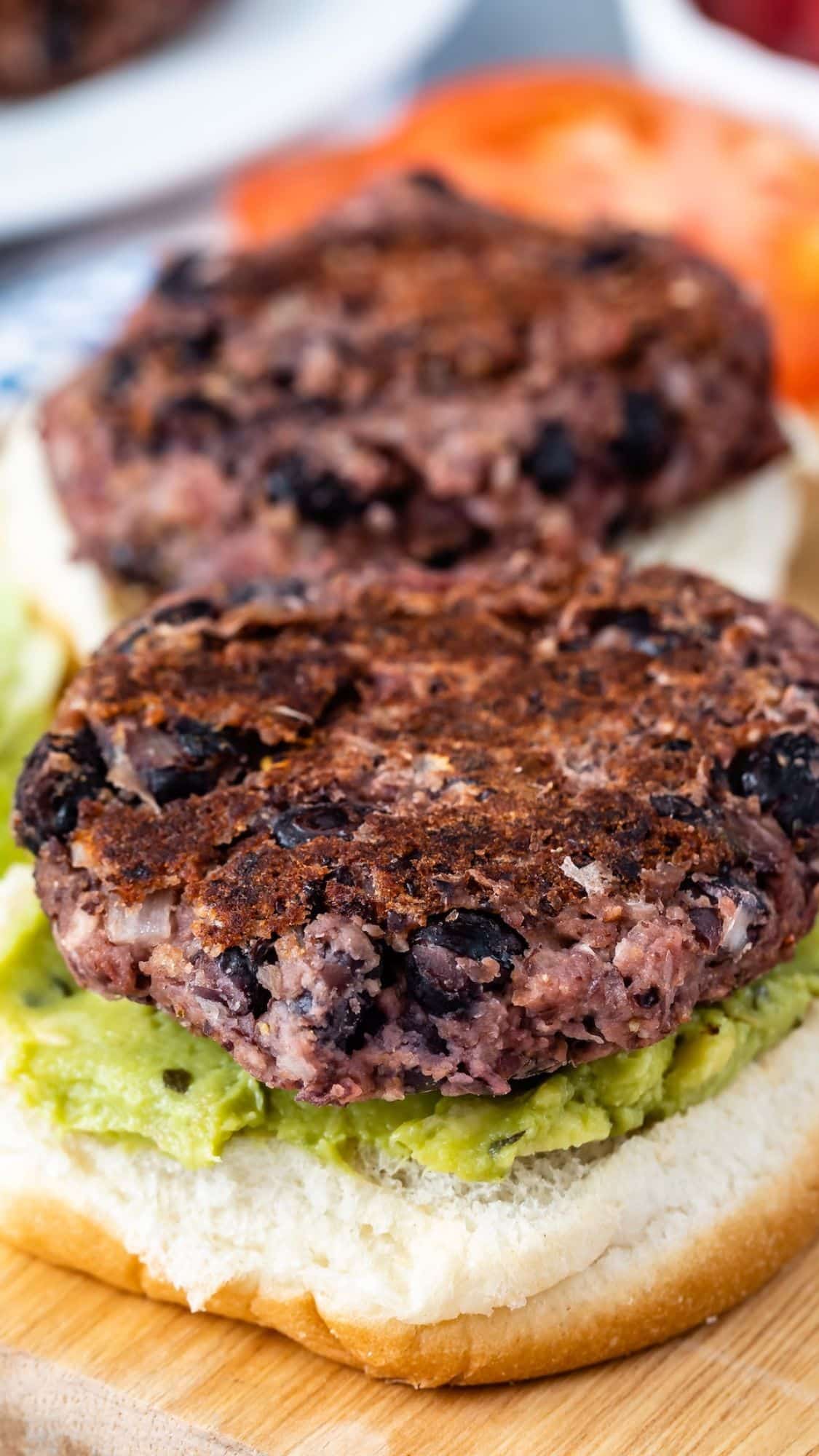A close-up of a black bean burger patty on a bun with mashed avocado. The patty is slightly charred, with visible black beans and grain texture. Slices of tomato are blurred in the background.