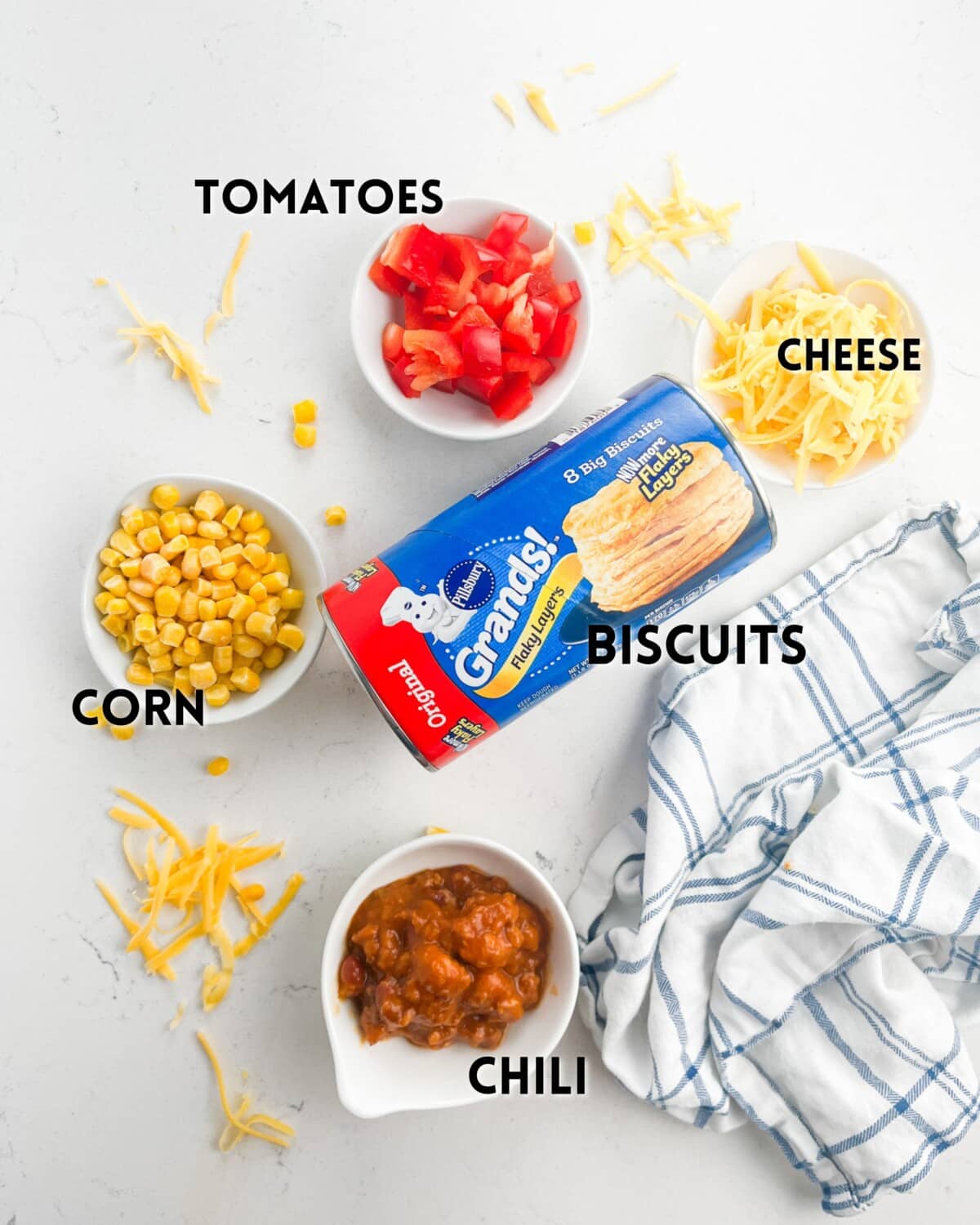 Top-down view of ingredients for a dish including a can of Pillsbury Grands biscuits, diced tomatoes, shredded cheese, corn, chili in a small bowl, and a striped cloth on a white surface.