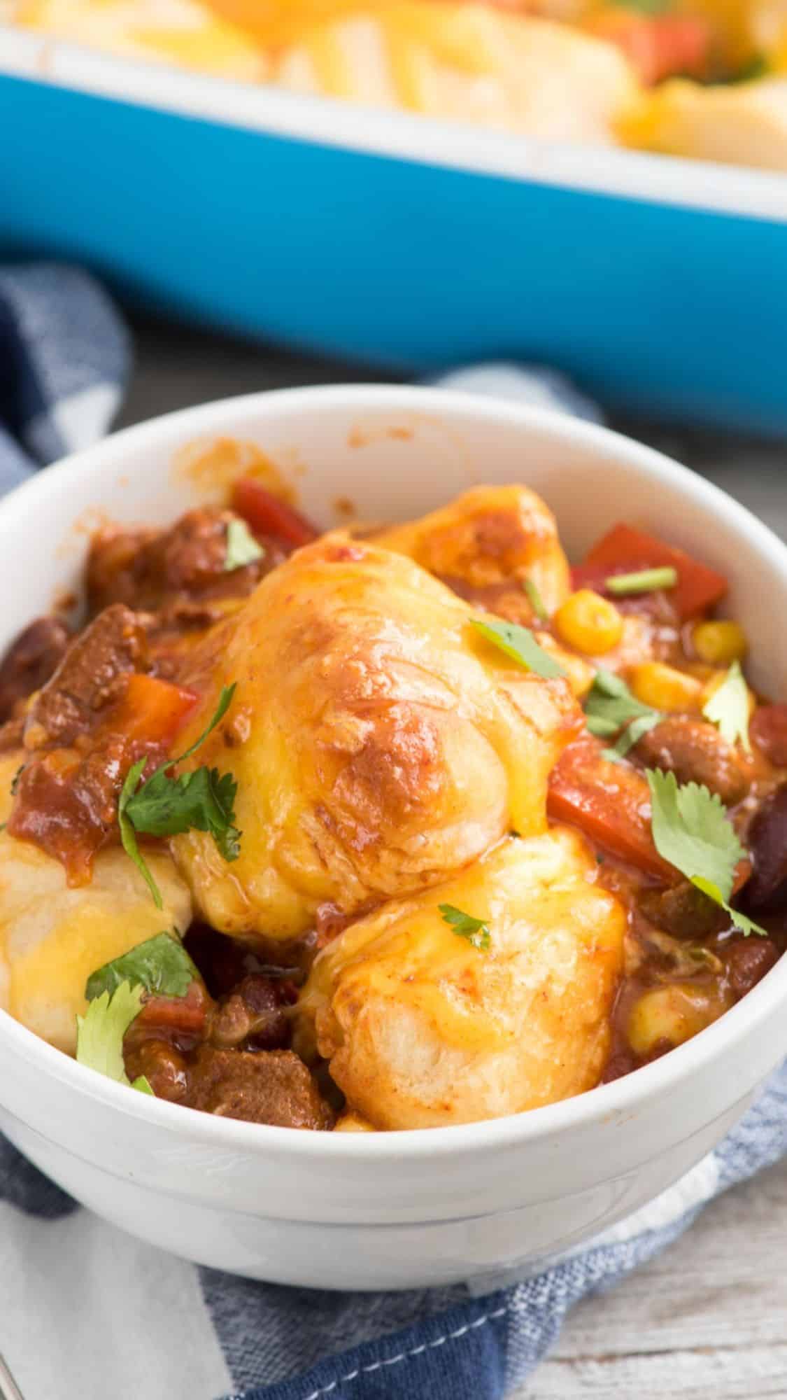 A bowl of cheesy chili, featuring chunks of beef, kidney beans, corn, and red bell peppers, topped with melted cheese and garnished with cilantro. A blue baking dish with similar contents is visible in the background.