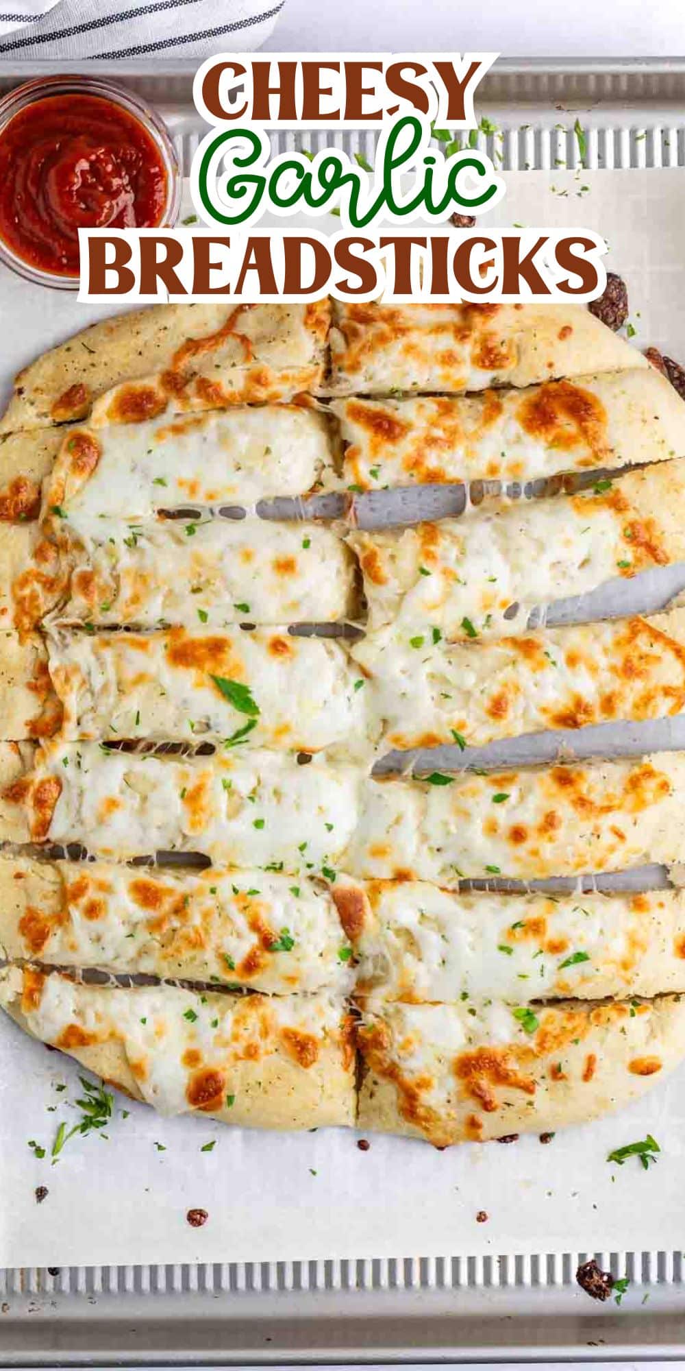 Cheesy garlic breadsticks topped with melted cheese and herbs on a baking tray. A small bowl of marinara sauce is visible in the corner. Text at the top reads Cheesy Garlic Breadsticks.