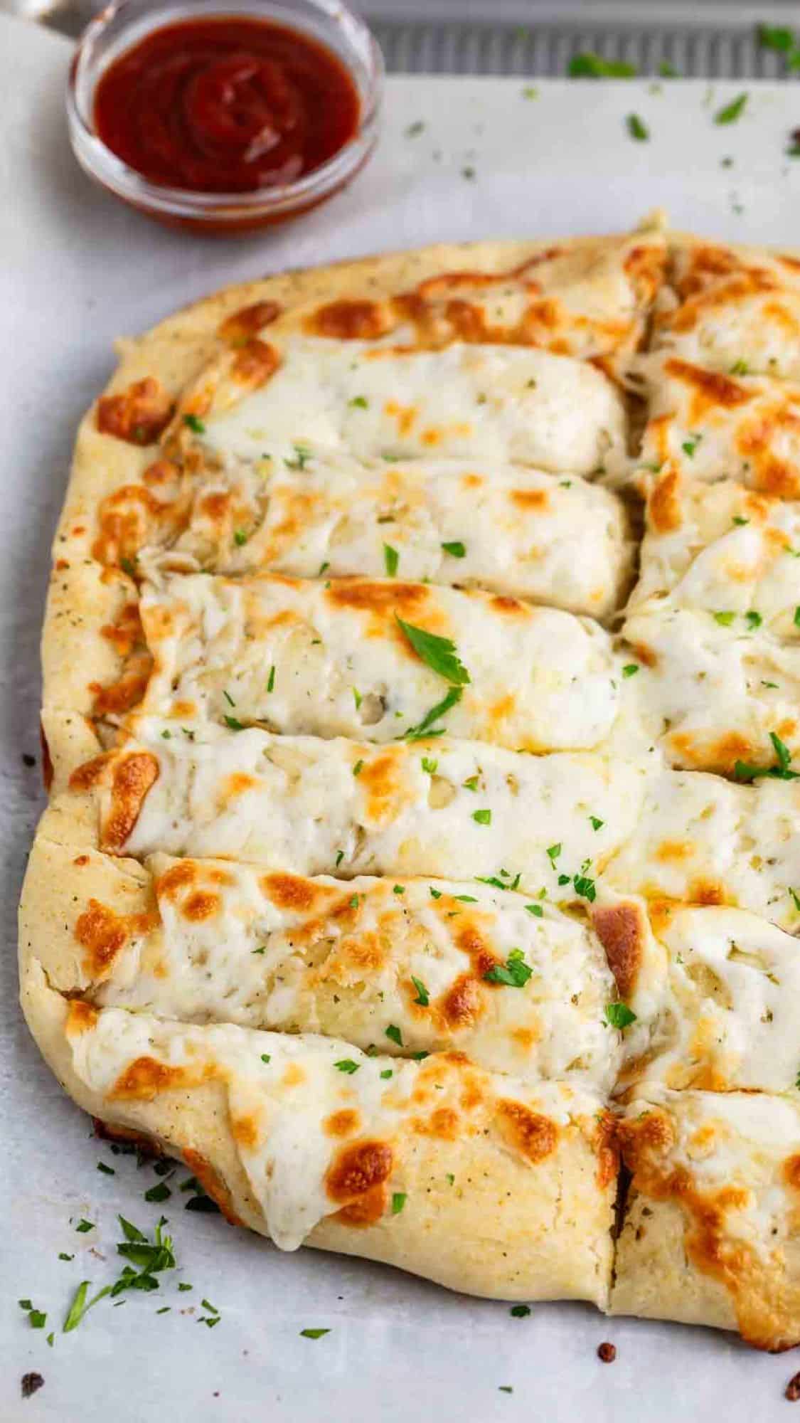 A rectangular cheesy garlic bread cut into strips, topped with melted cheese and sprinkled with herbs, lies on parchment paper. A small bowl of marinara sauce is in the background for dipping.