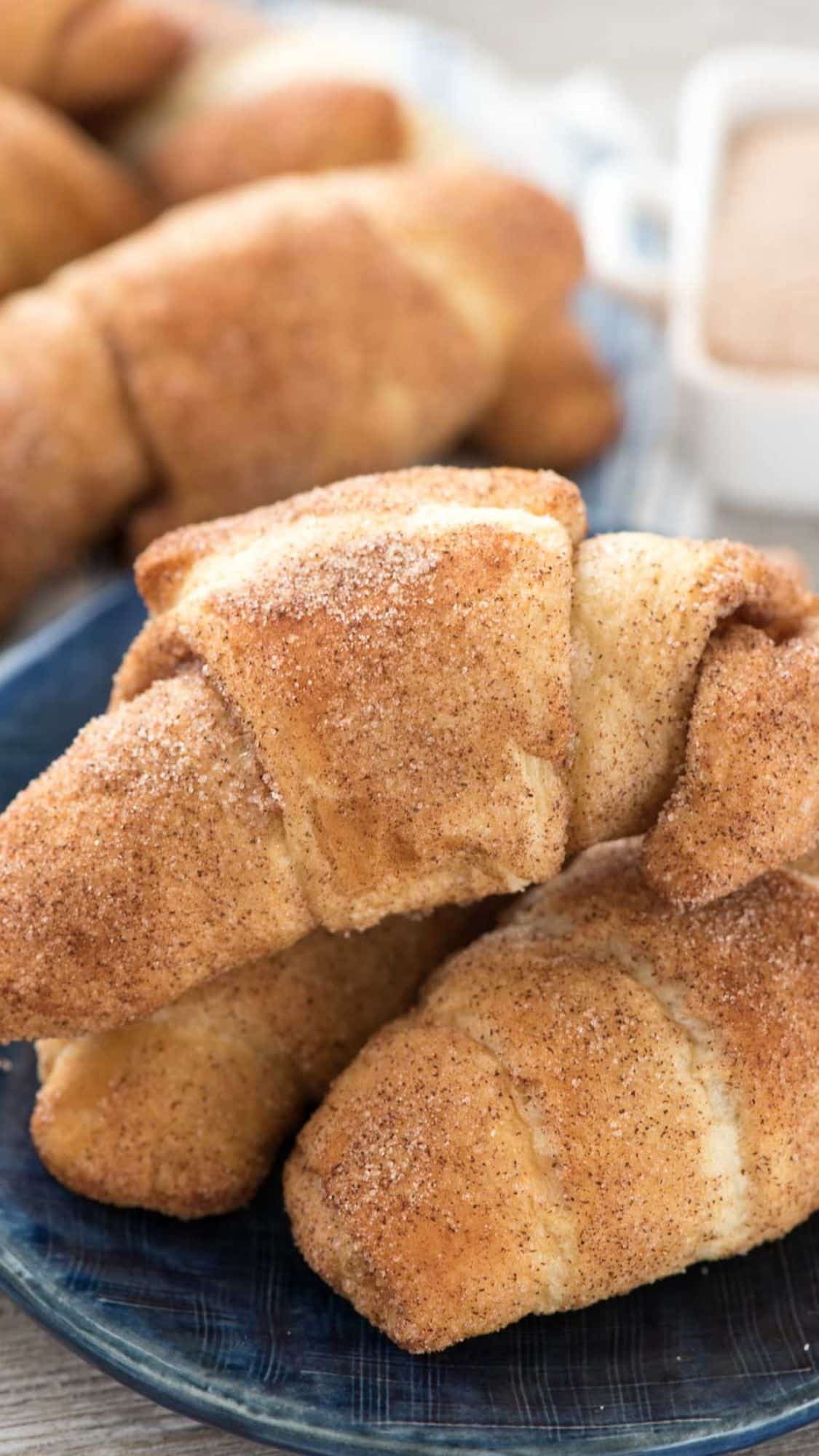 A plate of golden-brown crescent rolls covered in cinnamon sugar, stacked on a blue plate. A blurred container of spread is visible in the background.