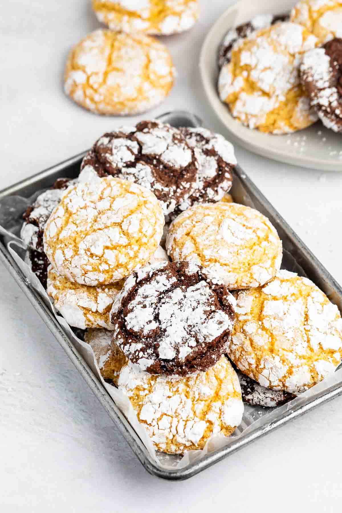A tray of yellow and chocolate crinkle cookies, dusted with powdered sugar. More cookies are seen on a plate in the background, all on a white surface.