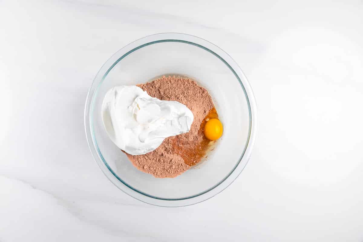 A glass bowl containing chocolate cake mix, a raw egg, and a scoop of whipped topping, all on a white countertop.