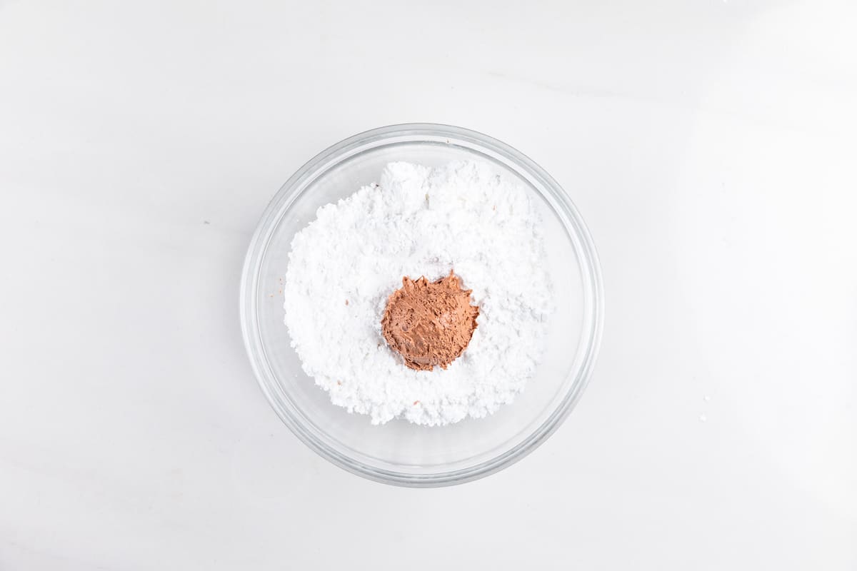 A glass bowl filled with white powdered sugar and a small mound of cocoa powder in the center, placed on a white surface.