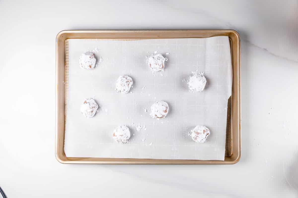 A baking sheet lined with parchment paper holds eight evenly spaced, round dough balls coated in powdered sugar, ready to be baked. The background is a white countertop.