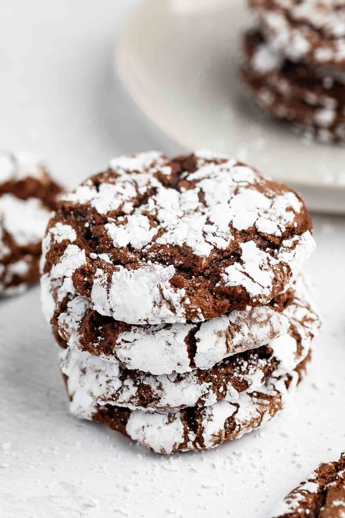 A stack of chocolate crinkle cookies coated in powdered sugar, with a plate of more cookies in the blurred background. The cookies have crackled surfaces, showing dark chocolate through the white sugar.