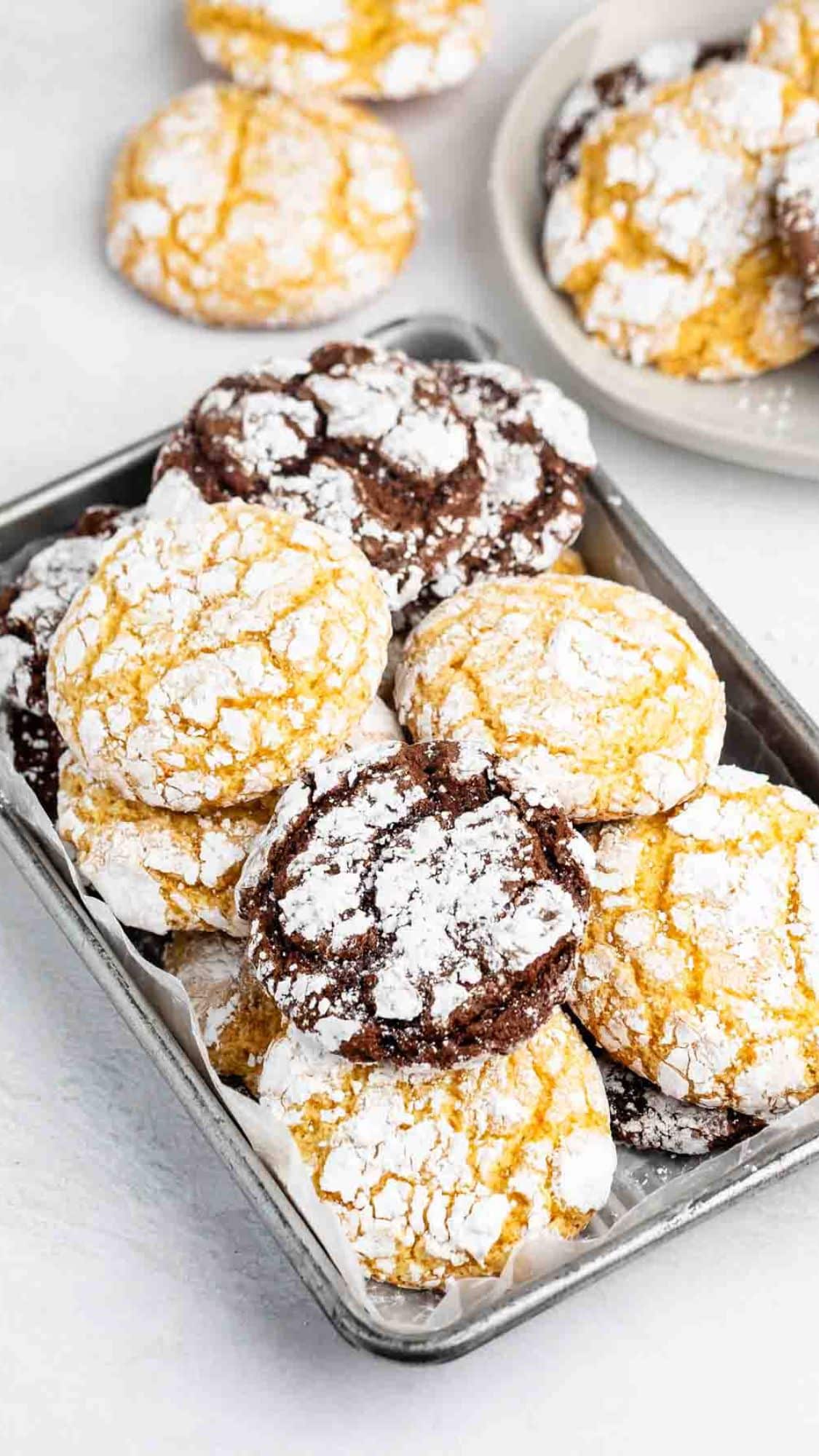 A tray filled with yellow and chocolate crinkle cookies dusted with powdered sugar, with more cookies on a plate in the background.